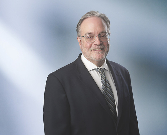 Man, Scott Chernich. wearing a dark blazer, standing against a blurred blue and white background.